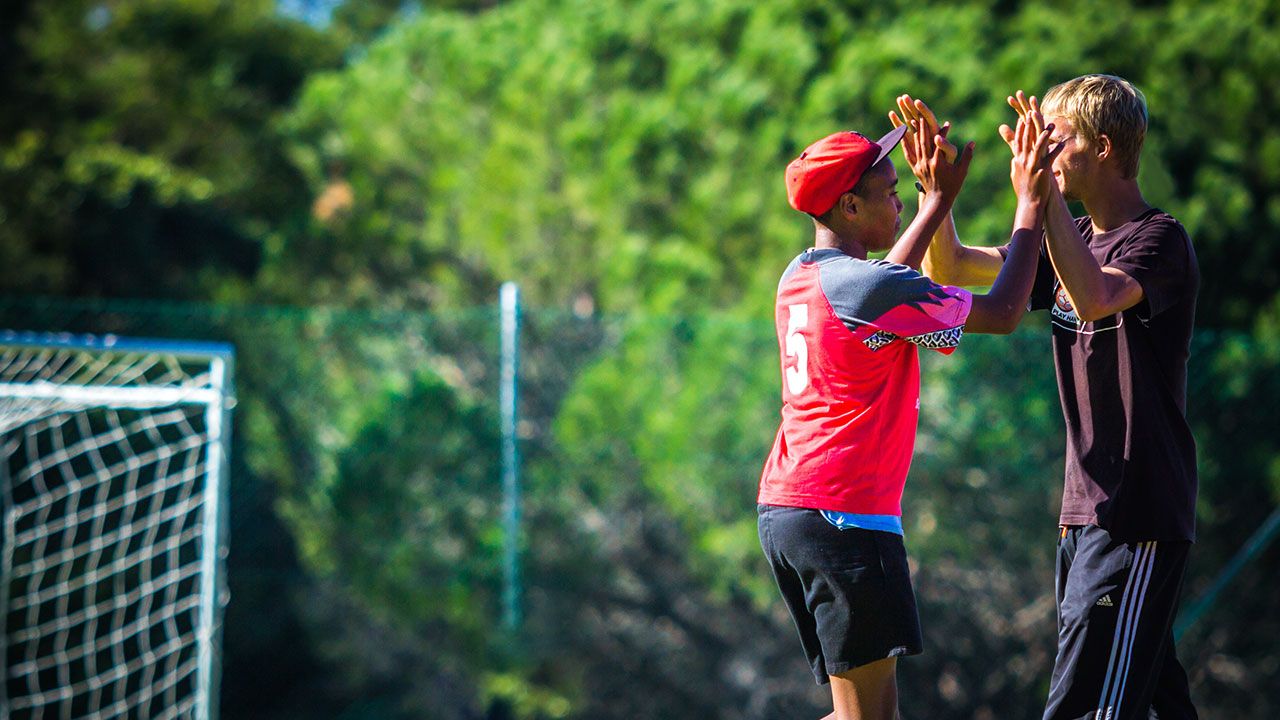 Auf dem Sportplatz eines sozialen Sportprojekts in Südafrika geben sich ein freiwilliger Sporttrainer und ein junger Spieler ein „High Five”.
